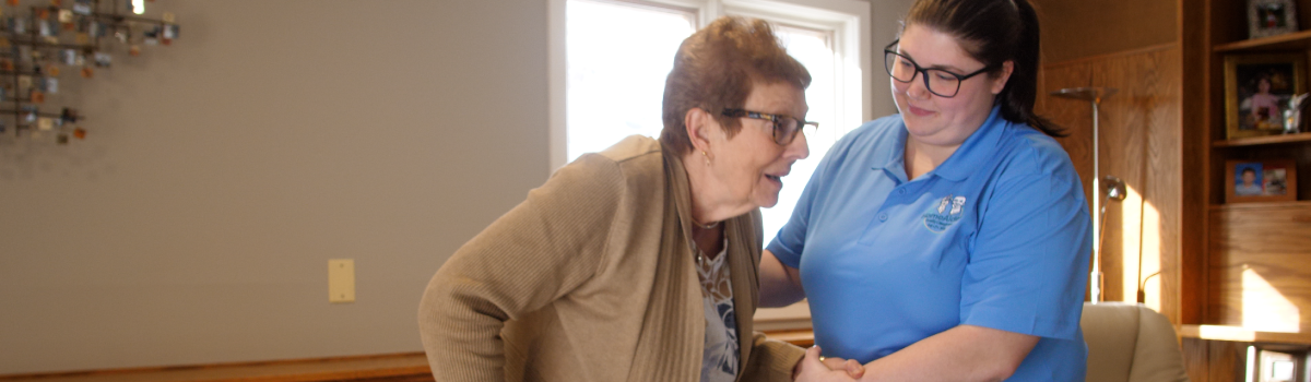 caregiver in a blue shirt helping an elderly woman walk throughout her home